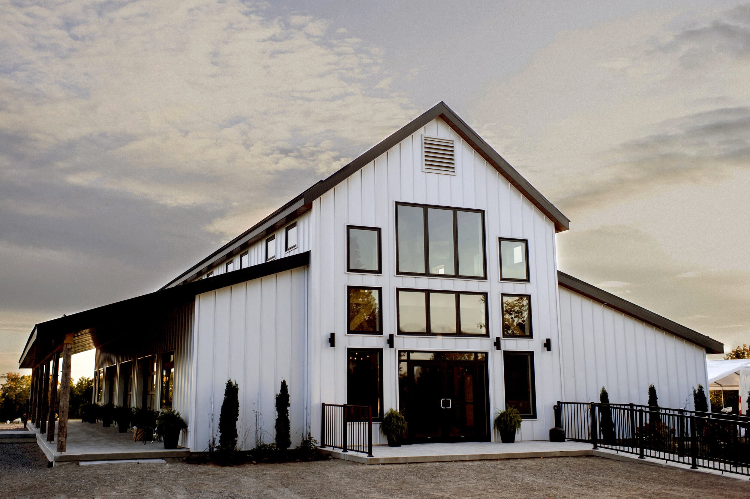 Exterior view of the modern white barn-style venue at Farmhill Weddings, featuring tall black-framed windows, a sloped roof, and rustic wooden pillars at sunset.